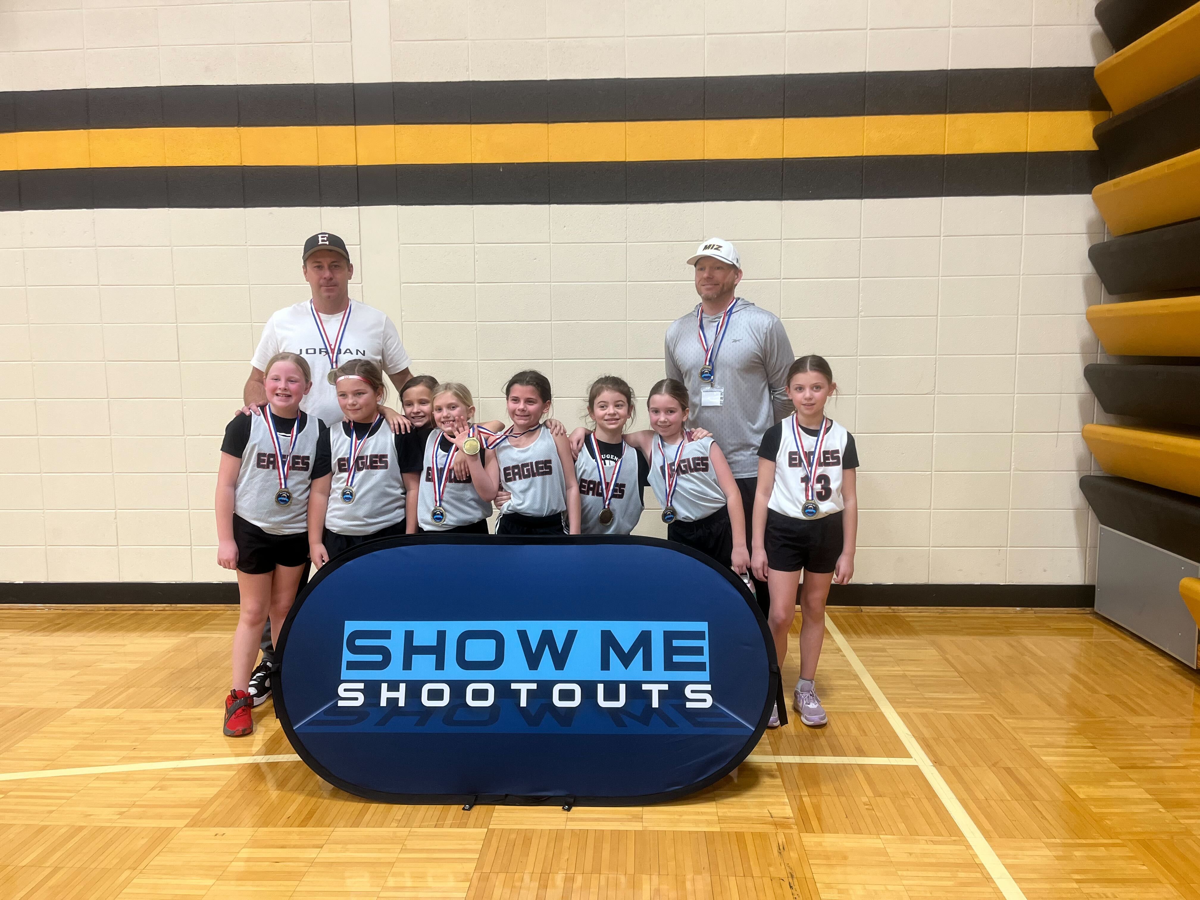 A young Eagles girls team and their coaches pose with medals behind the Show Me Shootouts banner.